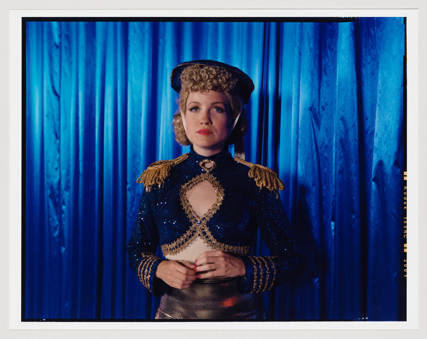 A woman in a blue sequined costume with gold epaulets stands before a blue curtain.