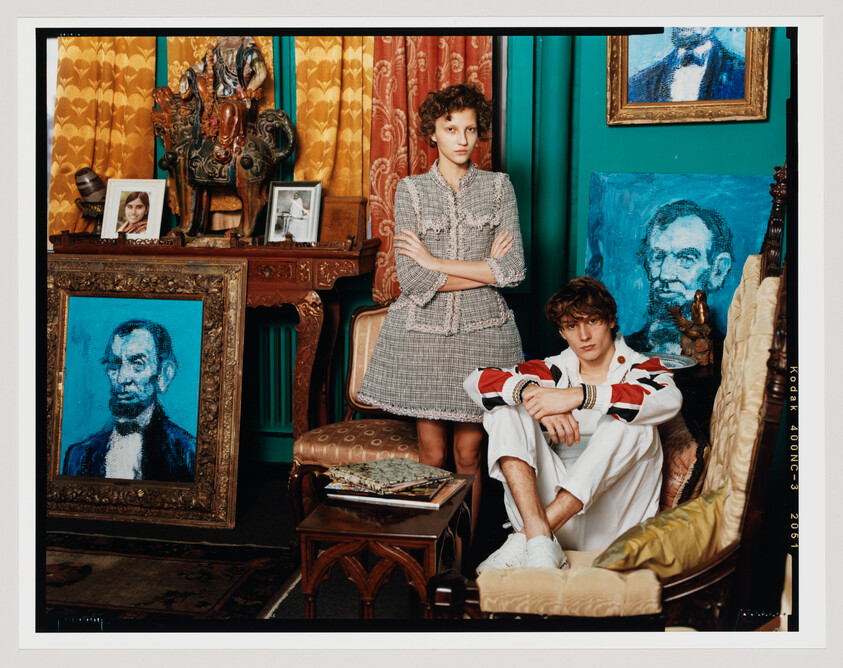 A stylized photograph of two individuals in a room filled with eclectic decor and artwork. The person on the left stands with arms crossed, wearing a tweed outfit, while the person on the right sits casually in an ornate chair, dressed in a white outfit with a red and black striped jacket. The room is adorned with various portraits, including blue-toned images of Abraham Lincoln, and is characterized by its vibrant curtains, patterned wallpaper, and antique furniture.