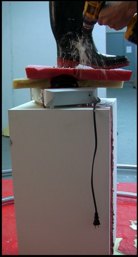 A person drills into a black boot standing on foam, causing liquid to spray out. An unplugged cord hangs nearby.