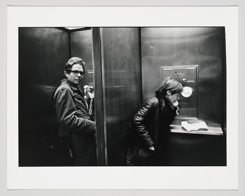A man and a woman use adjacent payphone booths, the woman speaking into the receiver while the man looks on.