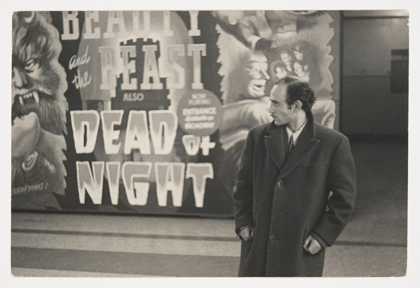 A man in a long coat stands looking to the side in front of a "Dead of Night" poster.