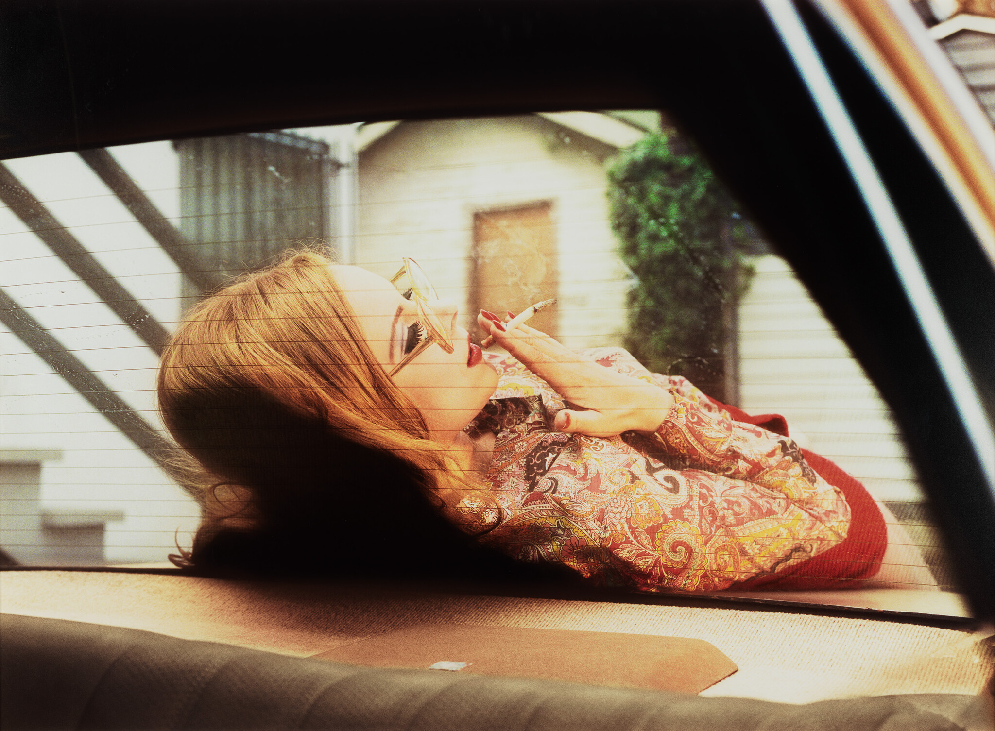 A woman wearing sunglasses lies across a car trunk smoking a cigarette.