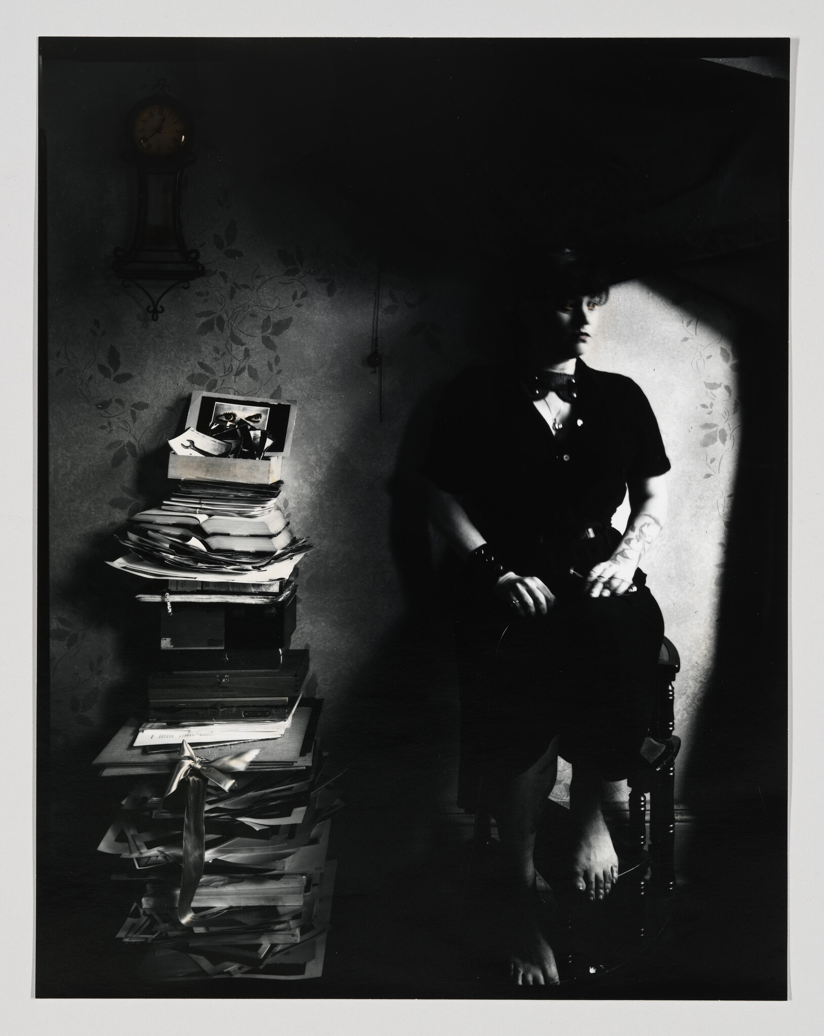 A person sits in shadow on a stool while a tall messy stack of papers stands beside them.