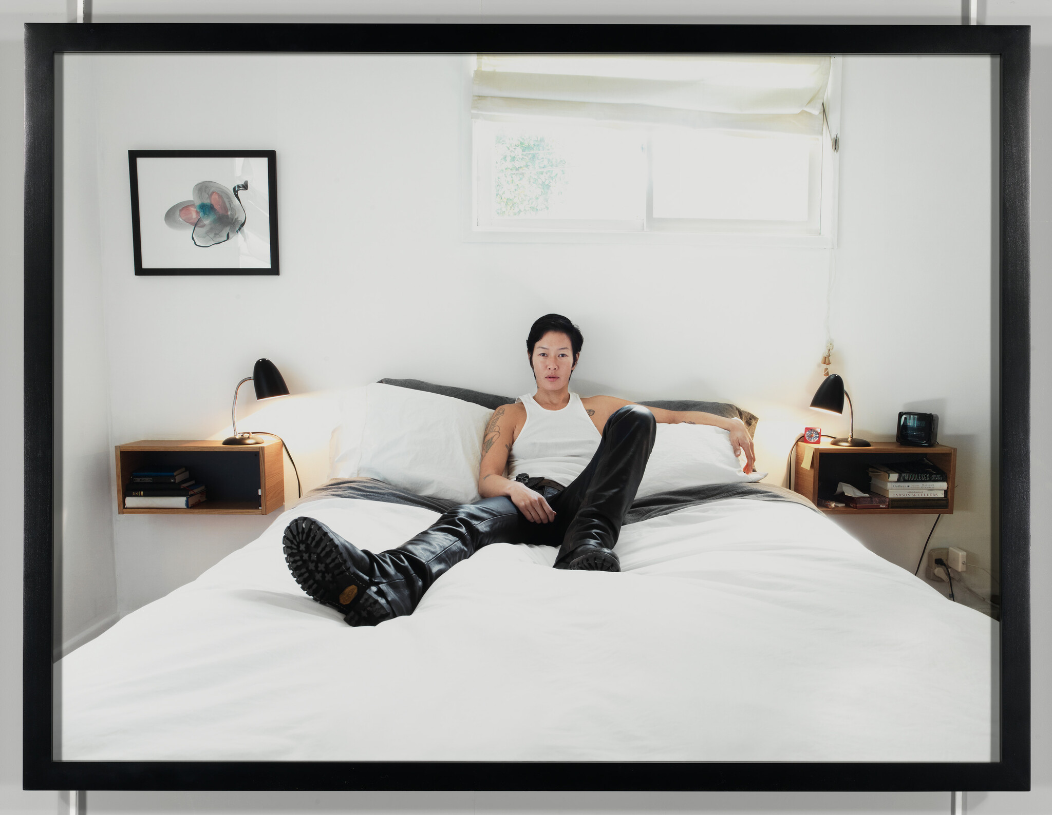A person wearing a white tank top and black leather pants with boots is reclining on a neatly made bed with white bedding. The room is minimalistic with two bedside tables, each with a lamp, and a framed picture on the wall. There is a window with a partially drawn blind in the background.