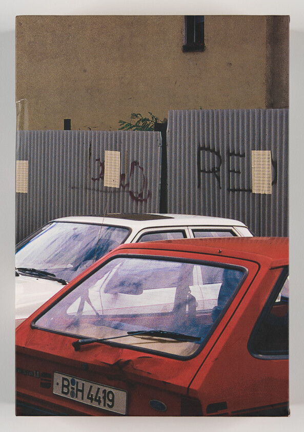 A red hatchback and a white car parked in front of a corrugated metal fence with graffiti.