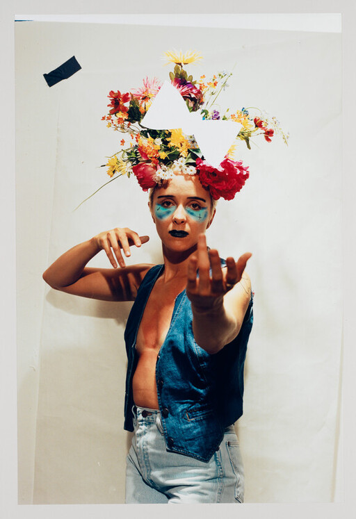 Person wearing a large floral headpiece and blue face paint reaches one hand toward the camera.