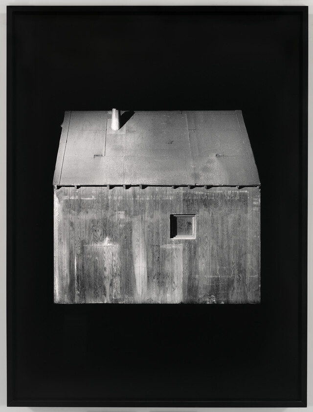 A black and white photograph of a simple, weathered wooden structure with a pitched roof and a small square window, displayed against a dark background within a frame.