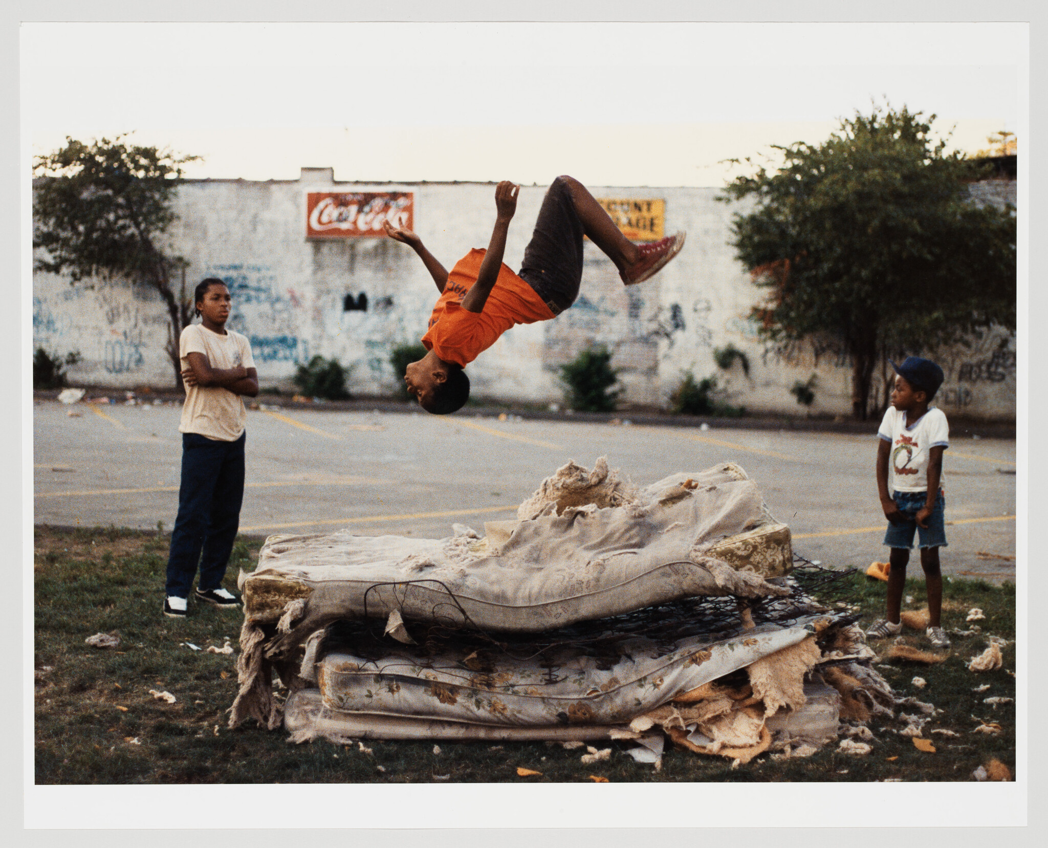 A boy flips backward onto a pile of old mattresses while two children watch nearby.