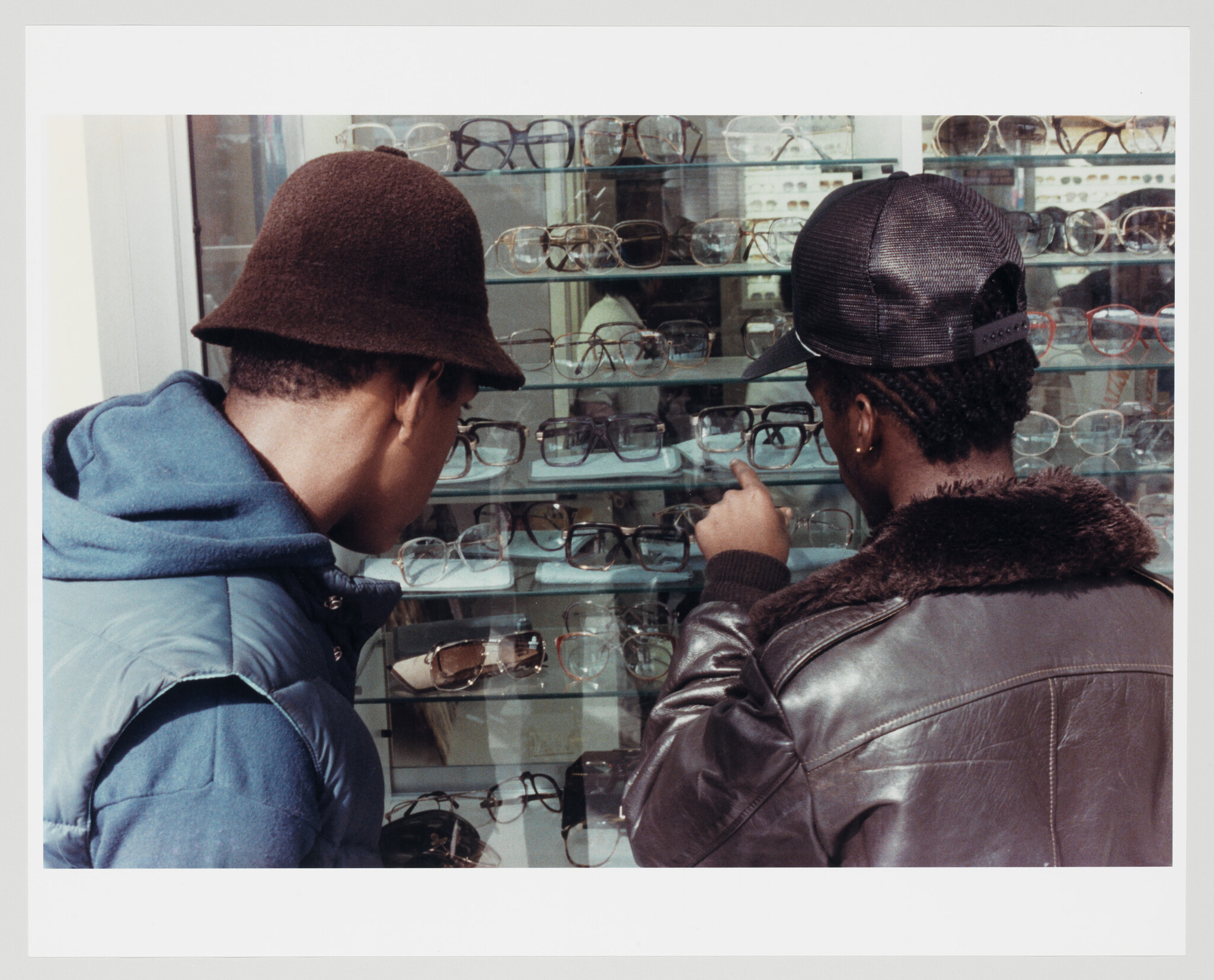 Two young men examine a shop window filled with eyeglasses while one points at a pair.