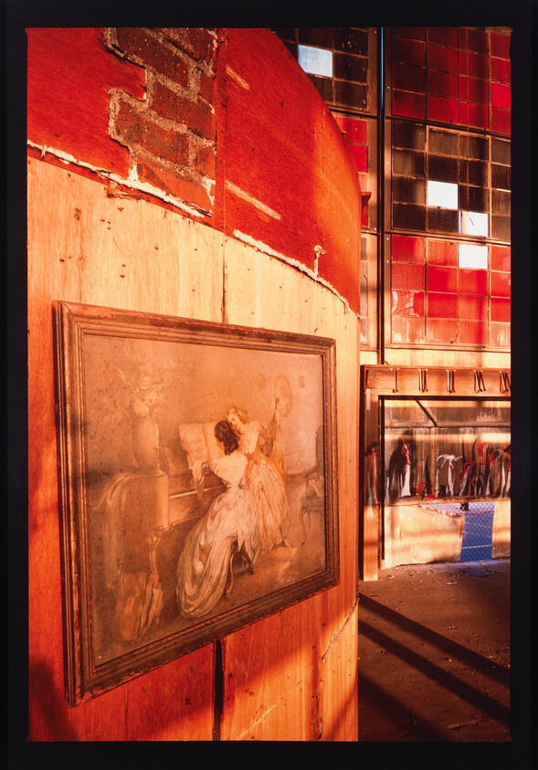 A dusty classical painting on a wooden wall inside a red-hued, abandoned building with broken windows reflecting clothing racks.