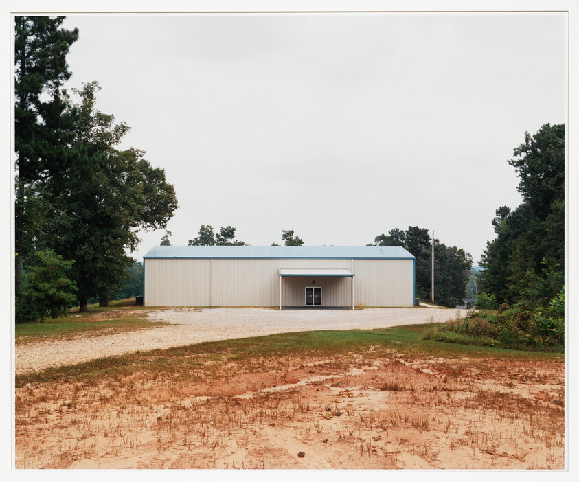 A small metal warehouse with a blue roof sits at the end of a gravel driveway.