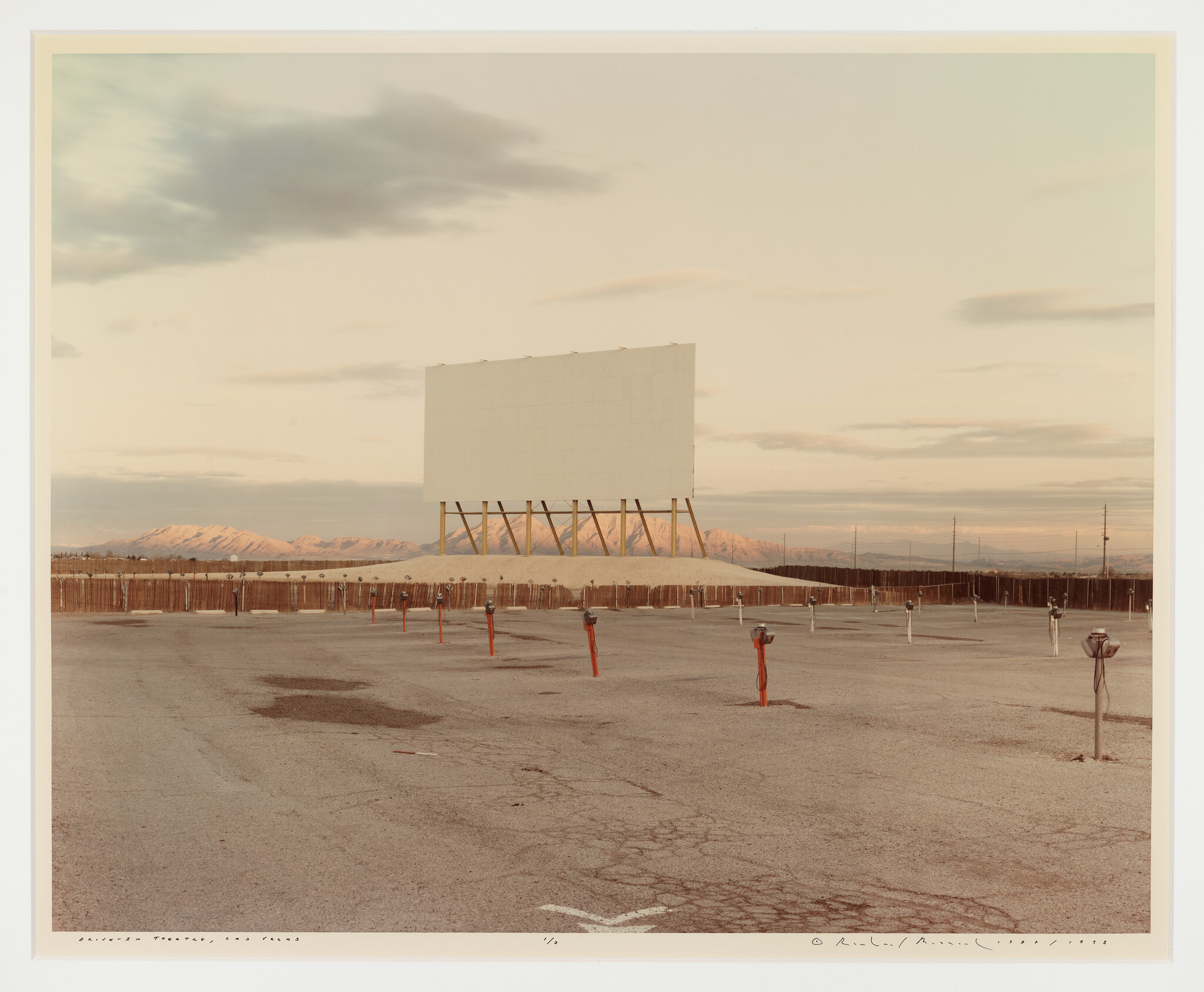 Large blank drive-in movie screen towers over empty parking lot with speaker posts and distant mountains.