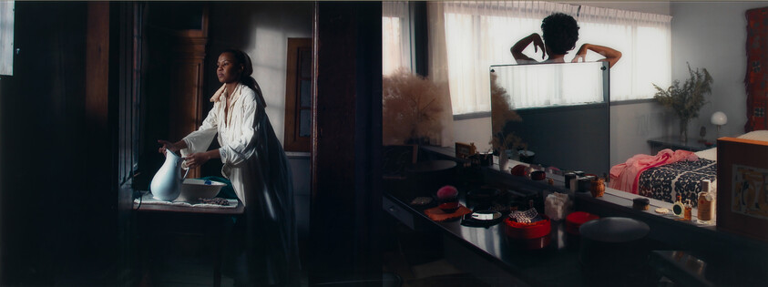 Two women in a bedroom area, one pouring water into a basin while another adjusts her hair at a mirror.
