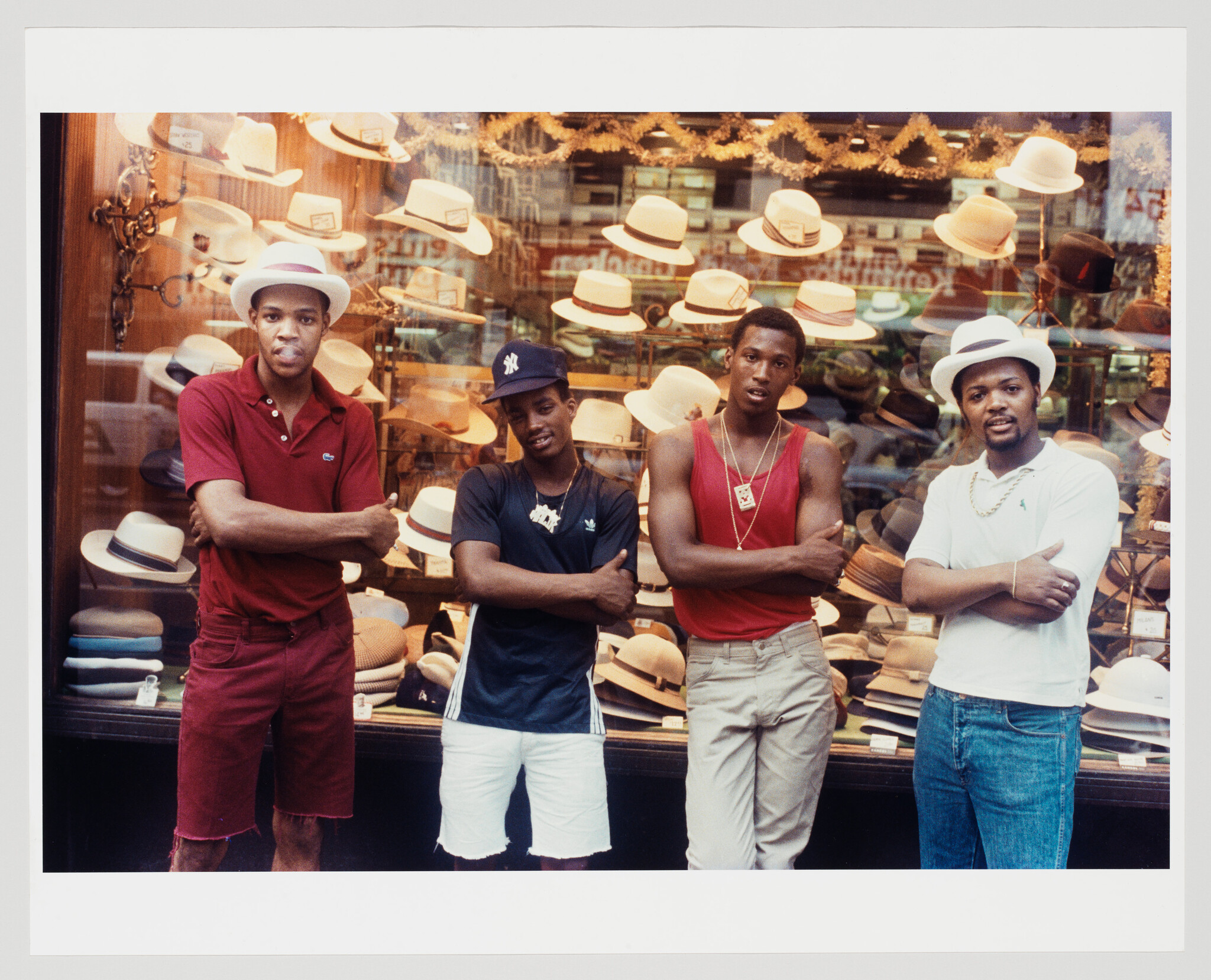 Four young men stand with arms crossed in front of a hat shop window.