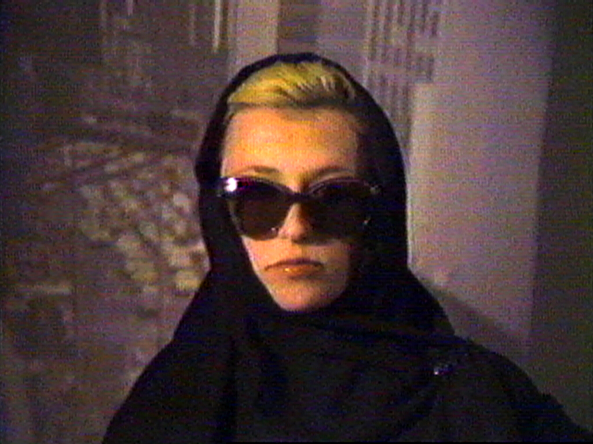 A person wearing a black headscarf and large sunglasses looks straight at the camera.