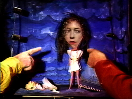 Two people point at a small doll with a woman's head and paper dress on a stage.