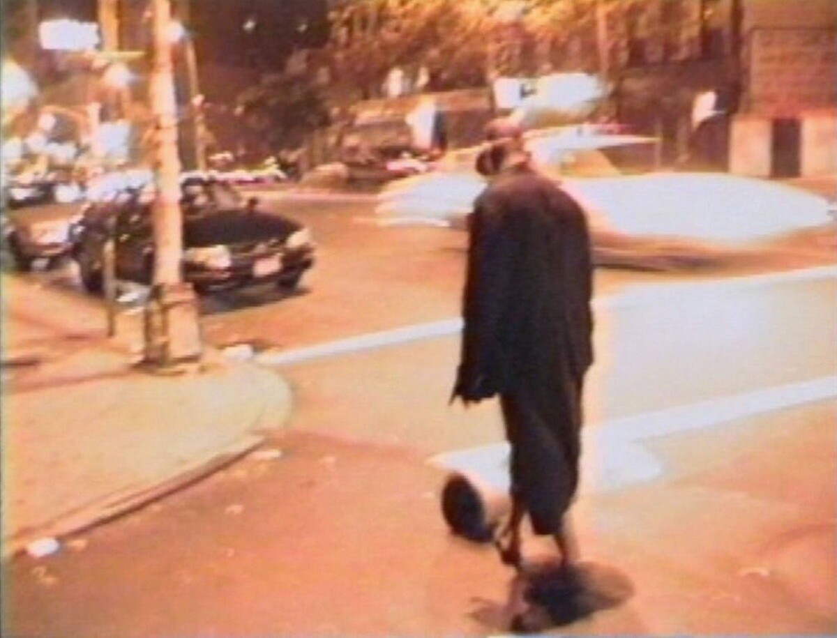 A lone person in a long coat walks across a dim city street at night.