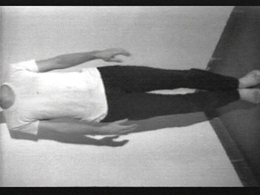 A person stands upright against a wall with feet together and arms relaxed at their sides.