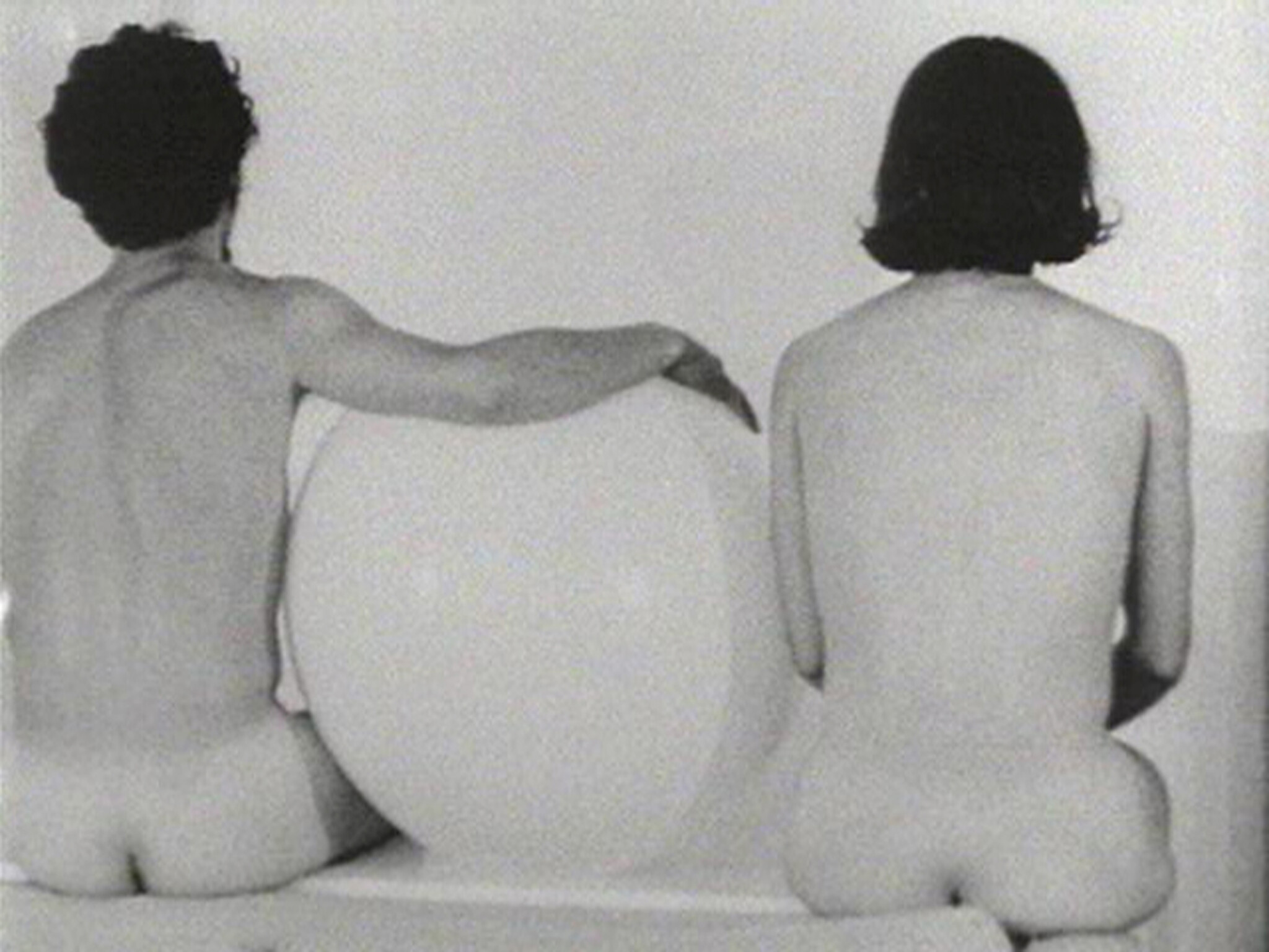 Two nude people sit side by side with their backs to the camera, separated by a large round object.