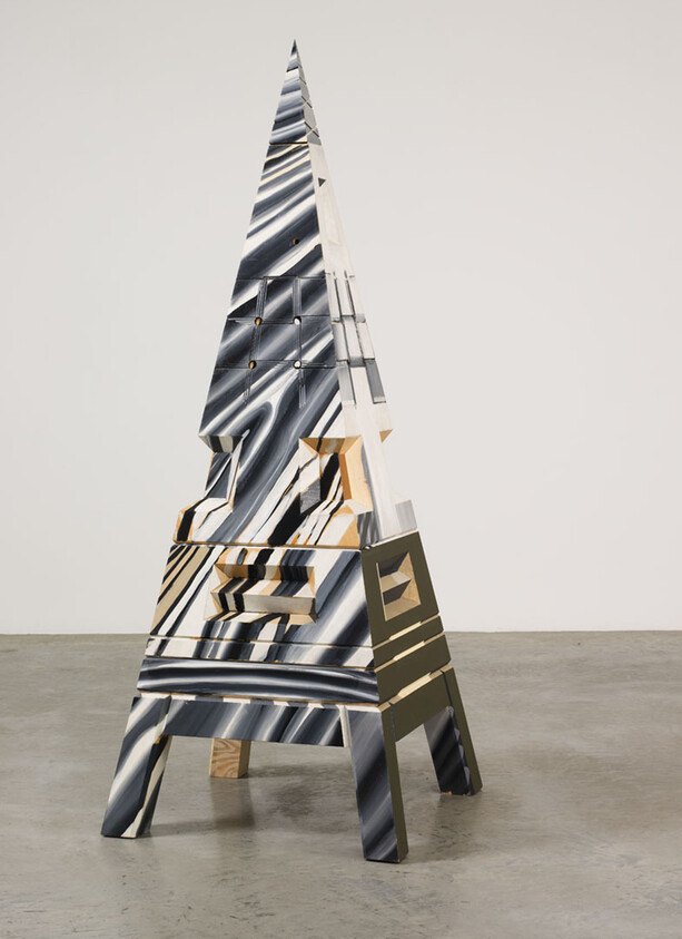 A modern, abstract sculpture resembling a distorted Eiffel Tower, with a metallic finish and geometric patterns, displayed in a gallery setting with a concrete floor.