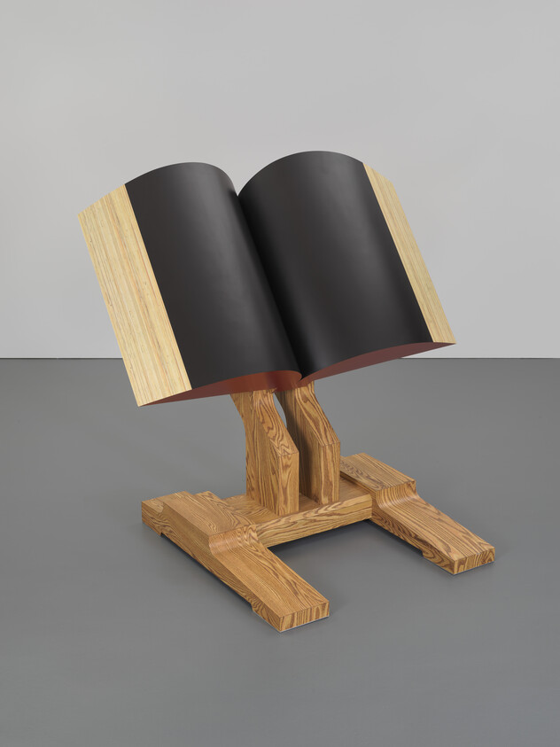 A sculpture resembling an open book with blank black pages, supported by a stylized wooden stand with a cross-shaped base, displayed against a grey background.