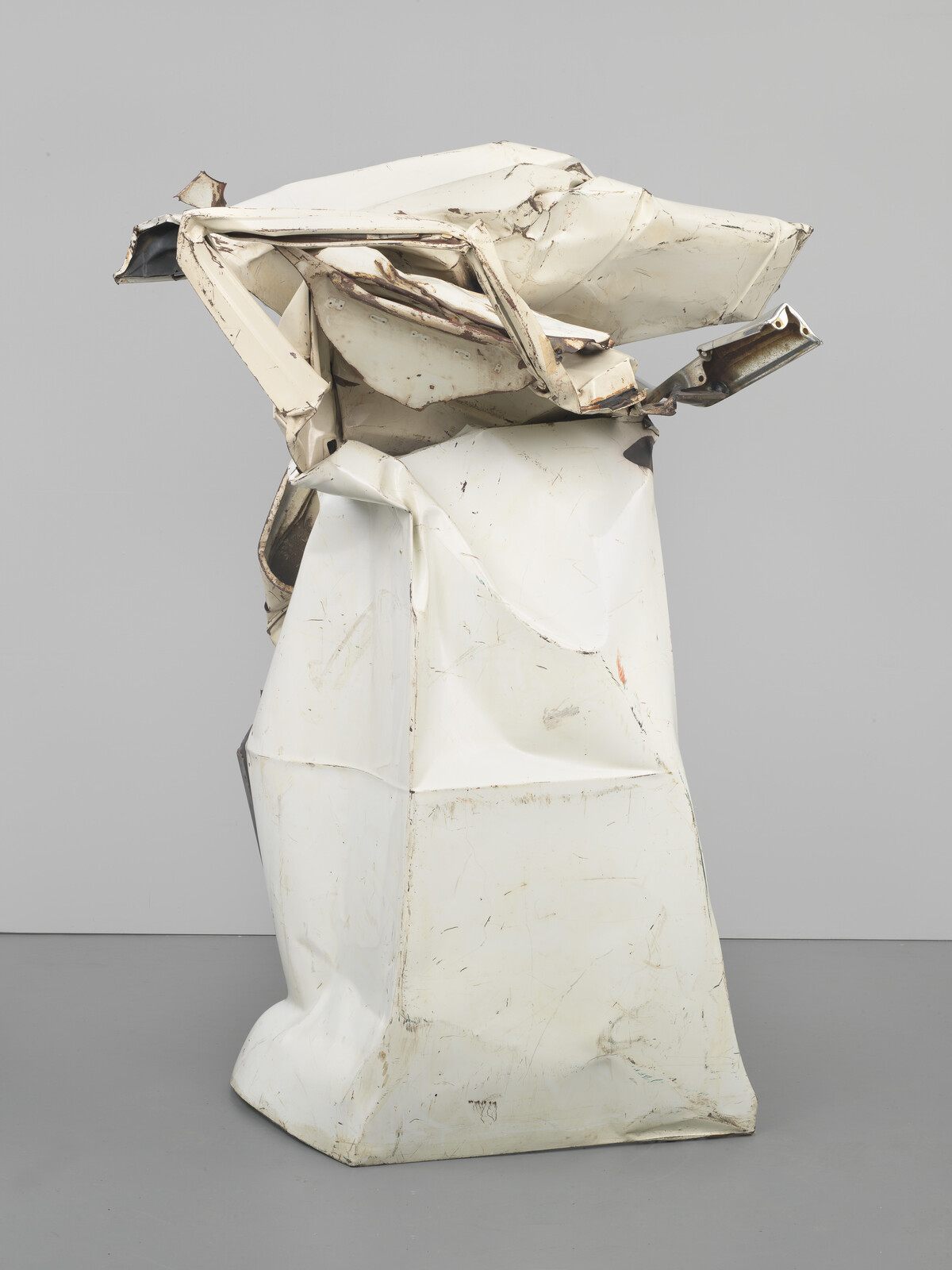 Tall crushed white metal sculpture with folded, dented panels standing against a plain gray background.