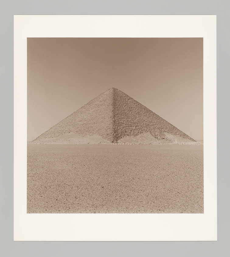 Sepia-toned photograph of a pyramid in a desert landscape, with a clear sky and sandy foreground.