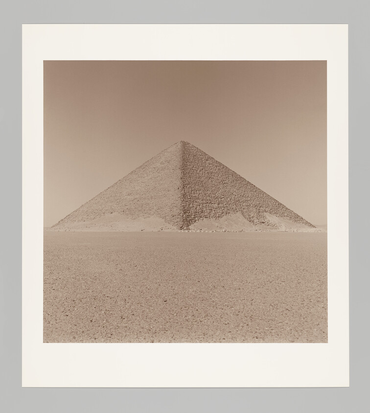 A lone ancient pyramid rises from a wide, flat sandy desert under a clear sky.