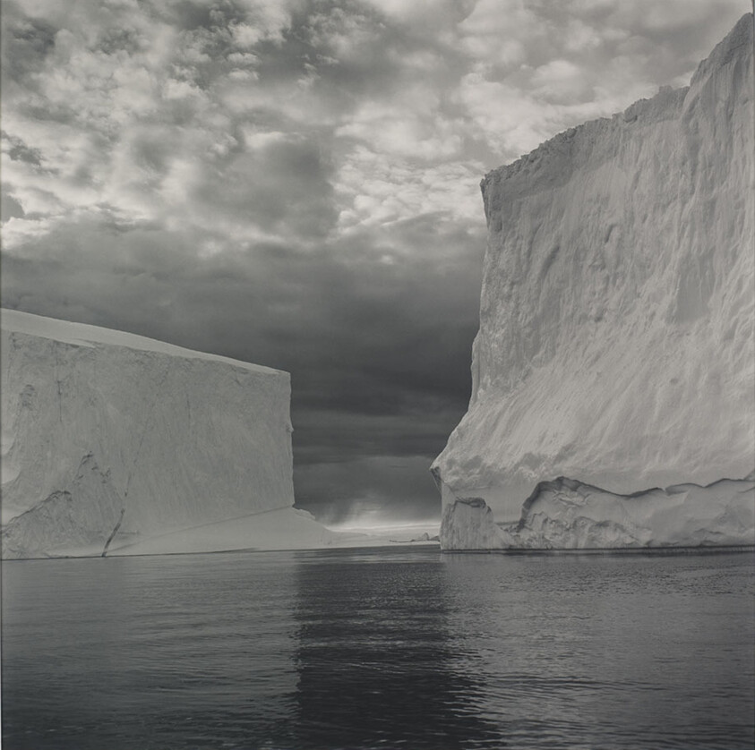 Two towering icebergs rise above calm dark water beneath a dramatic cloudy sky.