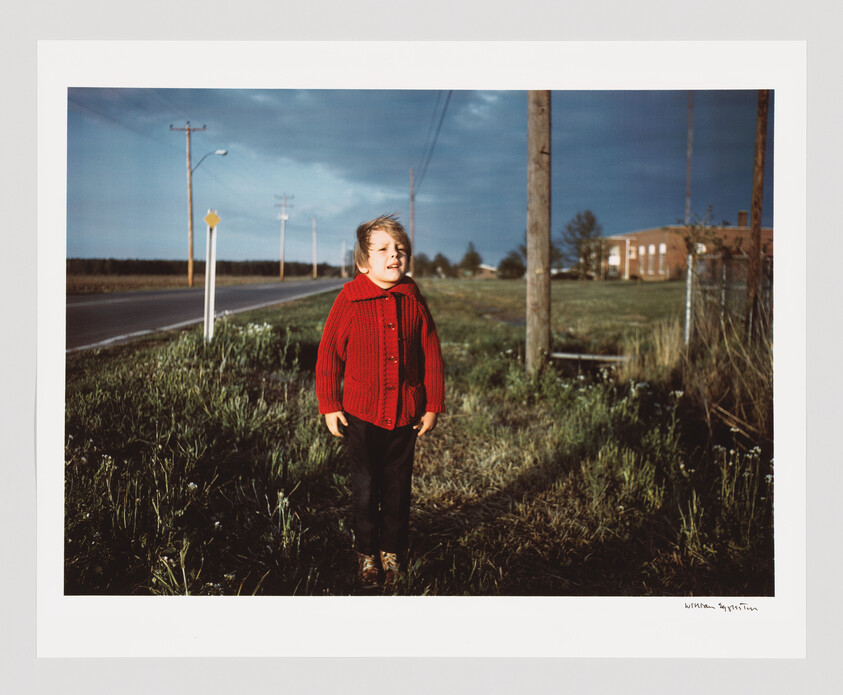 A young child in a red sweater stands by a rural road with wind tousling their hair.