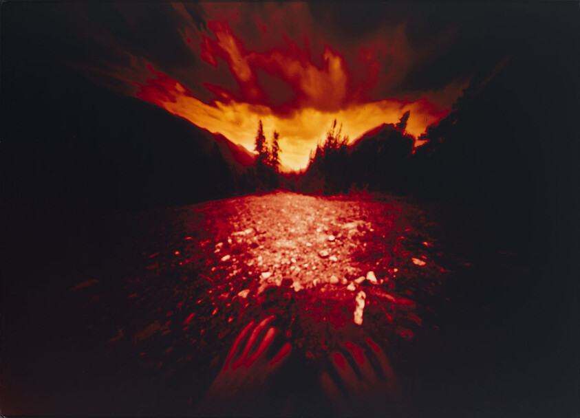 Someone's hands reach toward a rocky path under a dramatic red-orange sky at sunset.