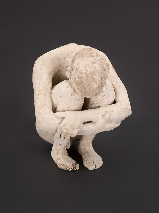 A white sculpture of a person crouching on a wooden floor, hugging their knees tightly.
