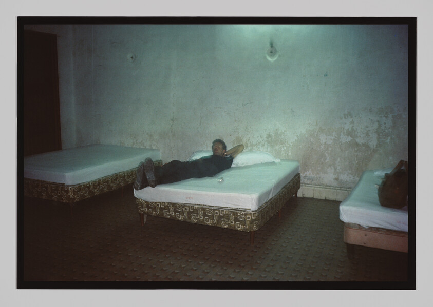 A person lies on a bed in a bare room with three beds and worn, peeling walls.
