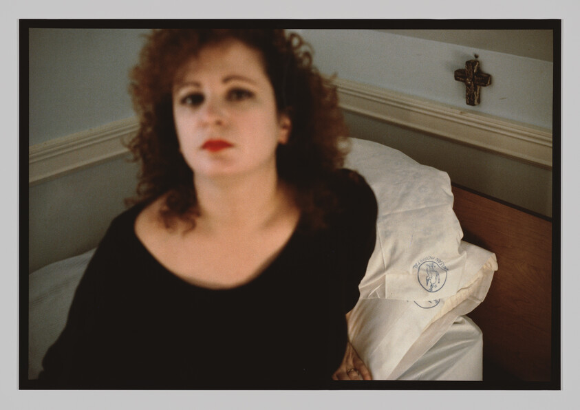 A woman with curly hair sits on a bed near white pillows, with a cross hanging on the wall.
