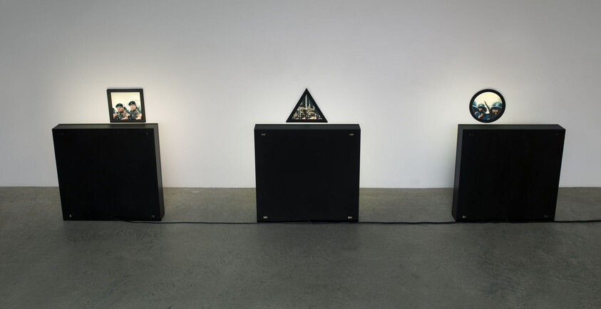Three black pedestals display small framed pictures in square, triangular, and circular shapes against a white wall.