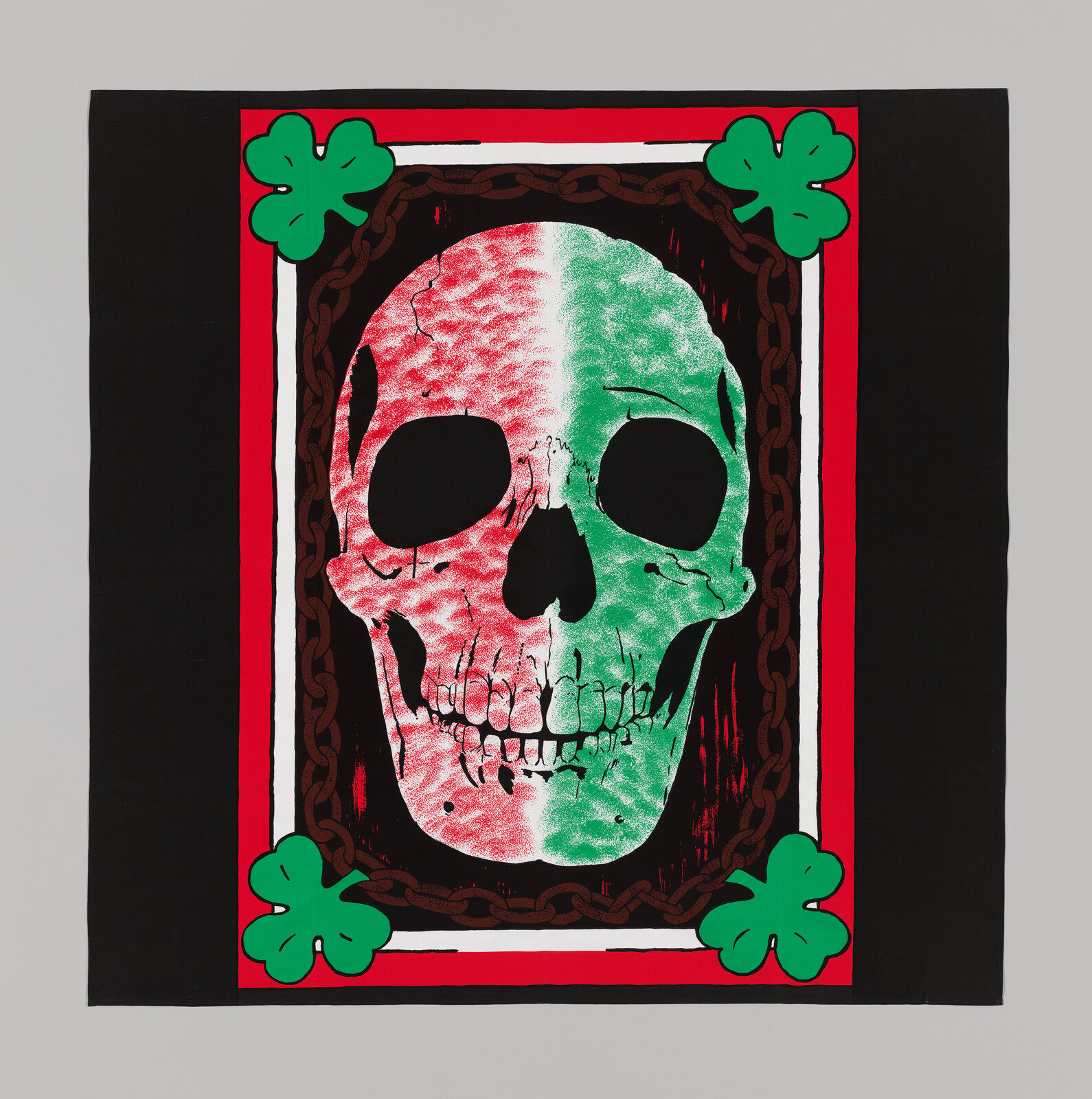 A large skull with red and green coloring is centered on a black cloth, bordered by four green clovers.
