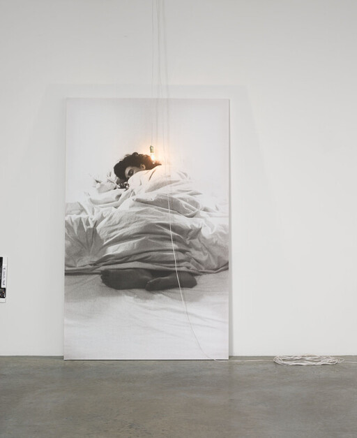 Large black-and-white portrait of a person sleeping under rumpled sheets with a hanging light.