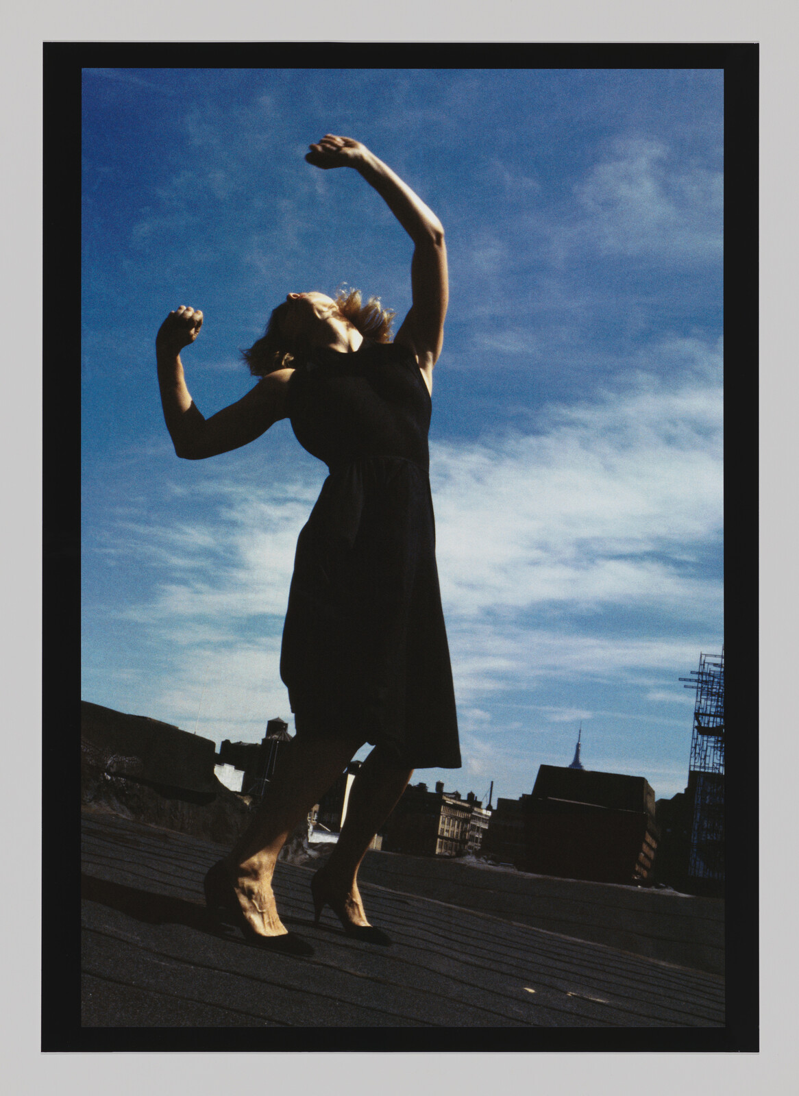 A woman in a black dress stands on a rooftop with arms raised against a blue sky.