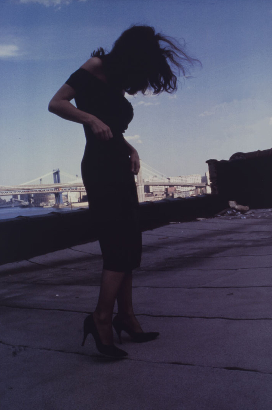 Woman in a black dress and high heels stands by the waterfront with hair blowing.