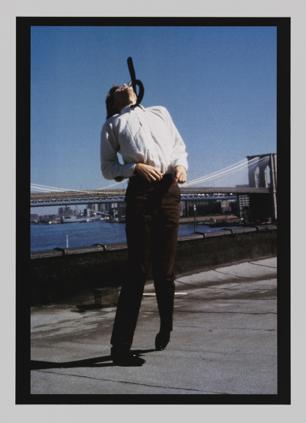 Person standing on a rooftop, leaning back with a black cane balanced on their open mouth. Bridges in background.