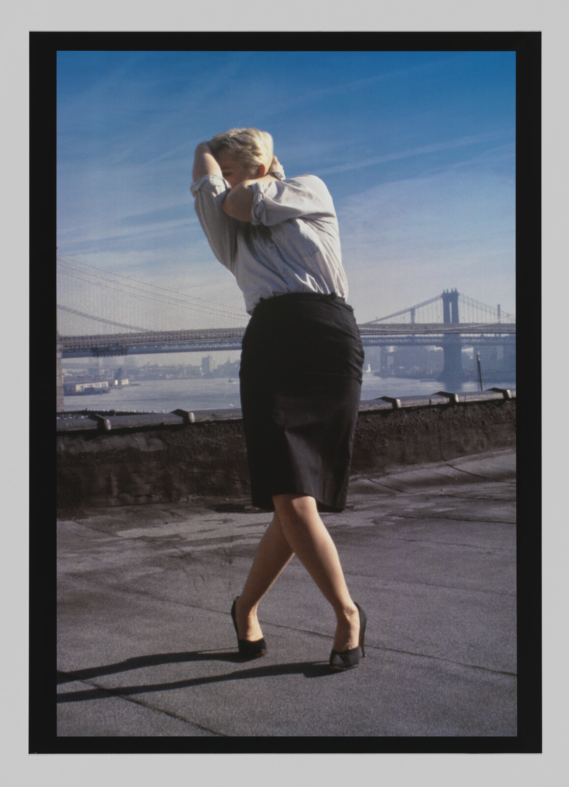A person in a skirt and heels stands on a rooftop with arms raised, bridges visible in the background.