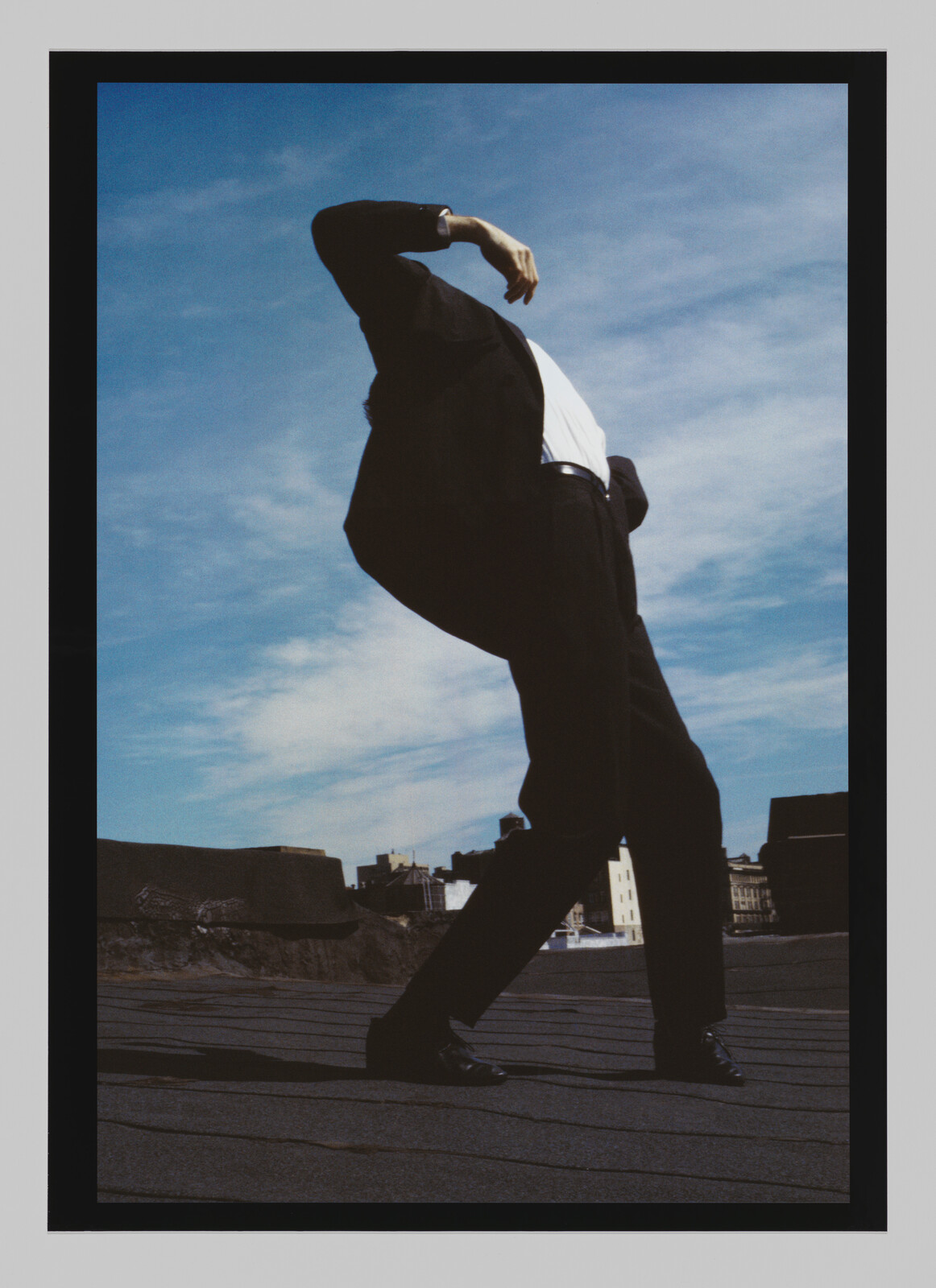 Person in a black suit and white shirt leans backward on a rooftop under a blue sky.