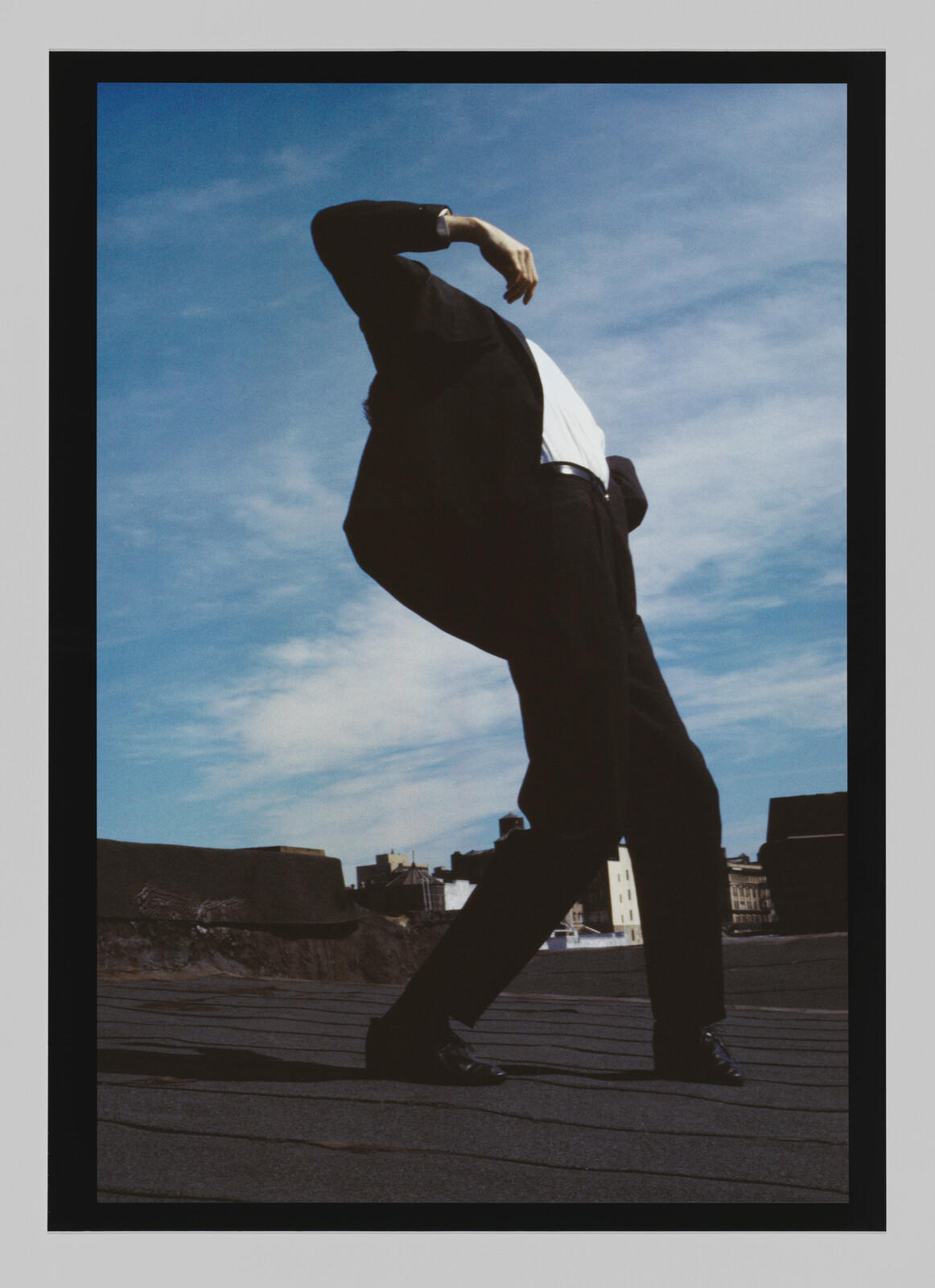 A person in a suit leans back dramatically on a rooftop against a blue sky.