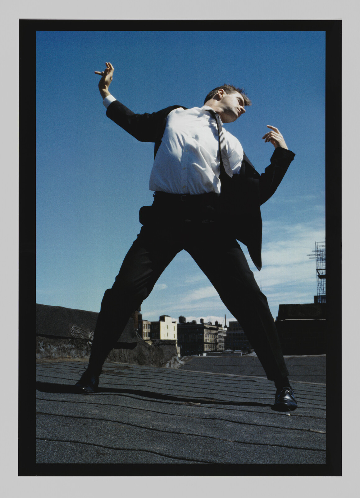 A person in a suit stands on a rooftop, leaning back with arms raised against a clear blue sky.