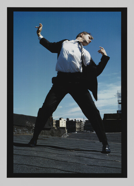 A man in a suit stands on a rooftop with legs apart, stretching arms toward the sky.