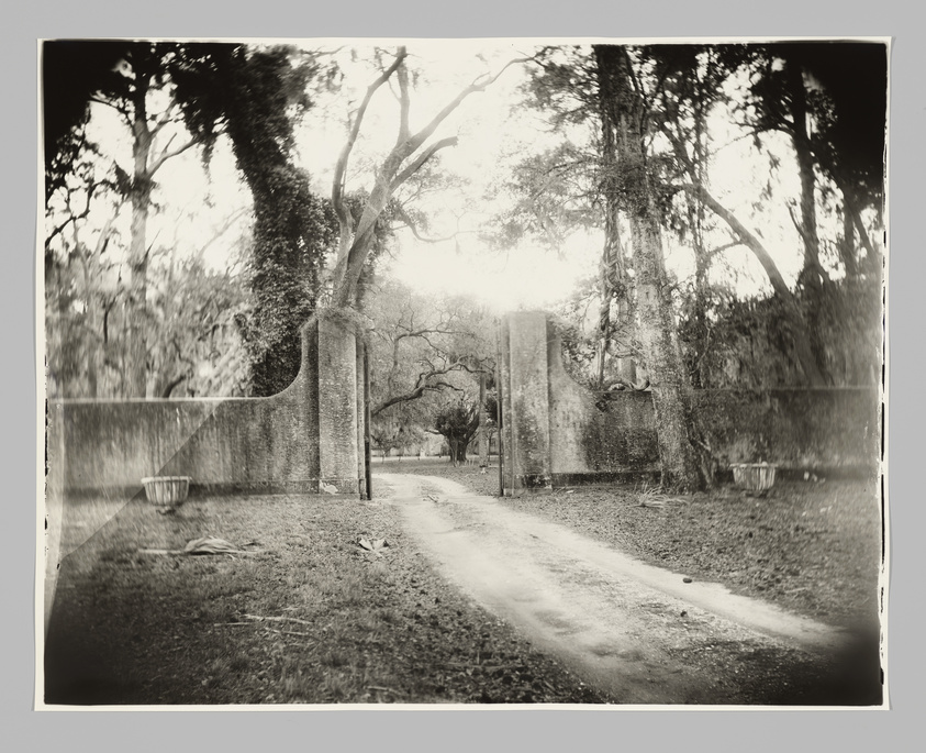 A dirt path leads through an open stone gate surrounded by tall trees, creating a serene and rustic atmosphere.