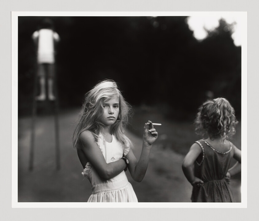 Young girl holding a cigarette, standing with two other children in a dimly lit outdoor setting.