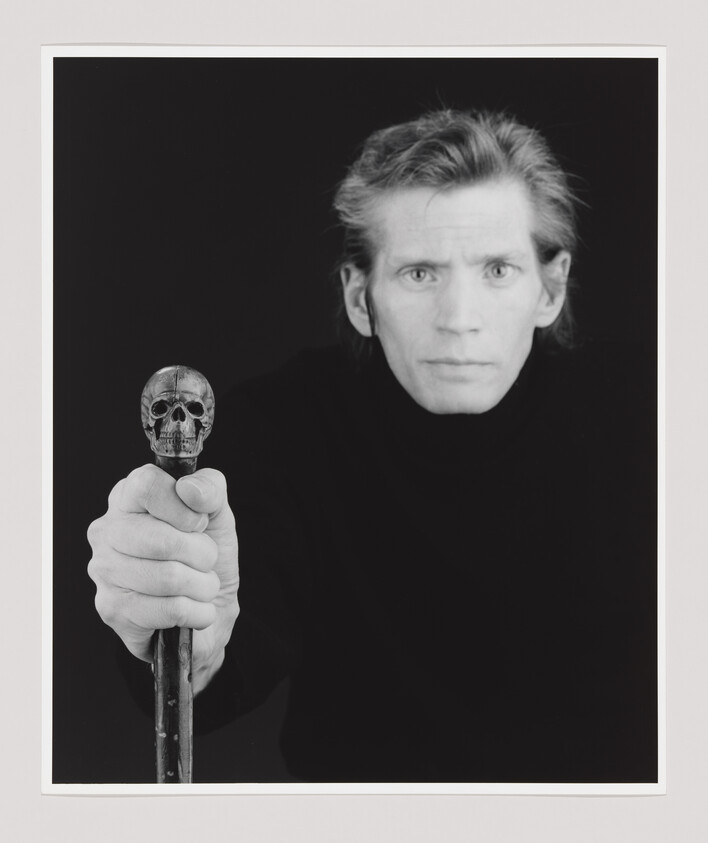 A person with light hair and a serious expression holds a cane topped with a skull toward the camera.