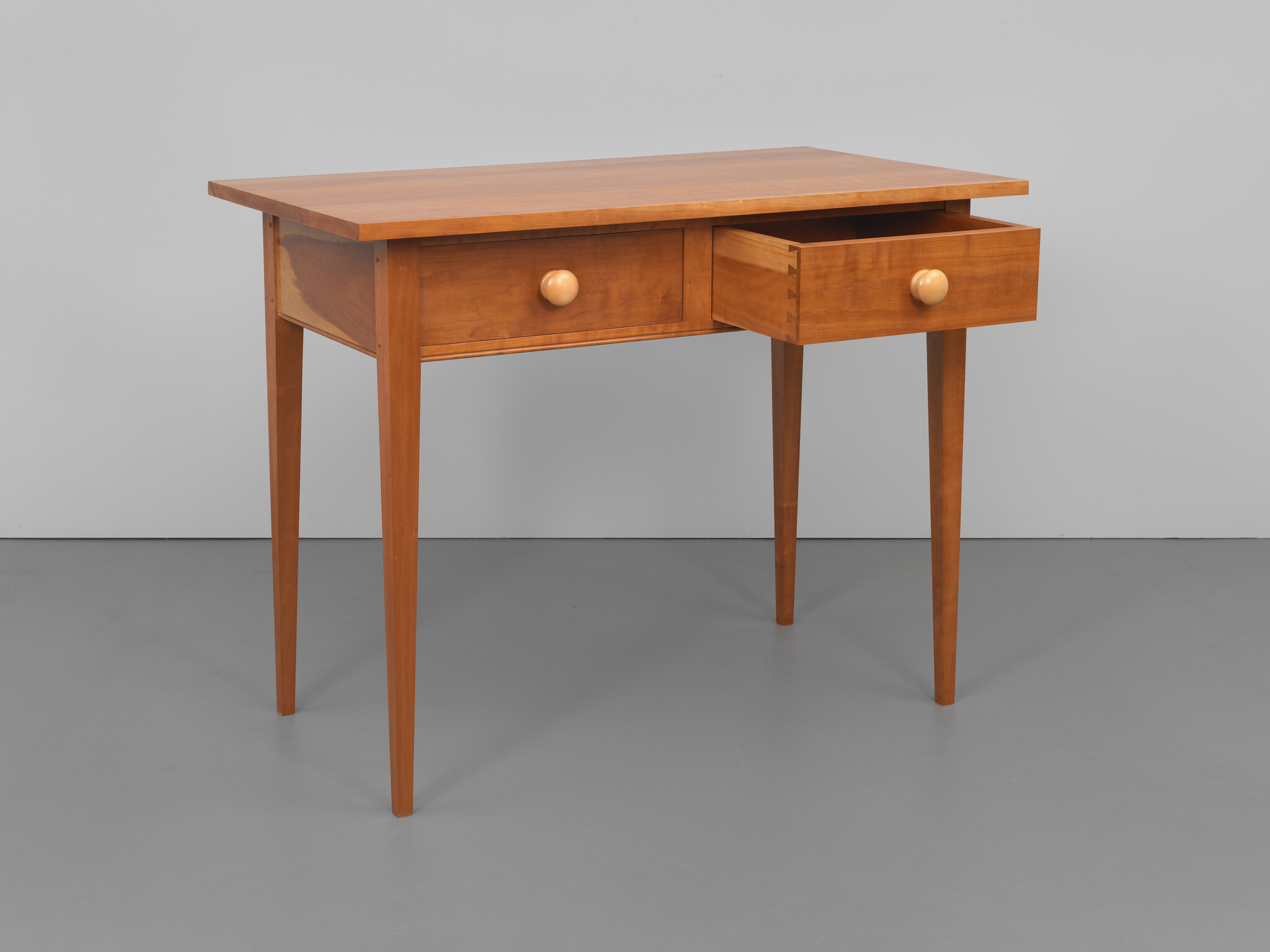 A wooden writing desk with a smooth finish, featuring two drawers with round knobs, standing on four slender, straight legs against a grey background.