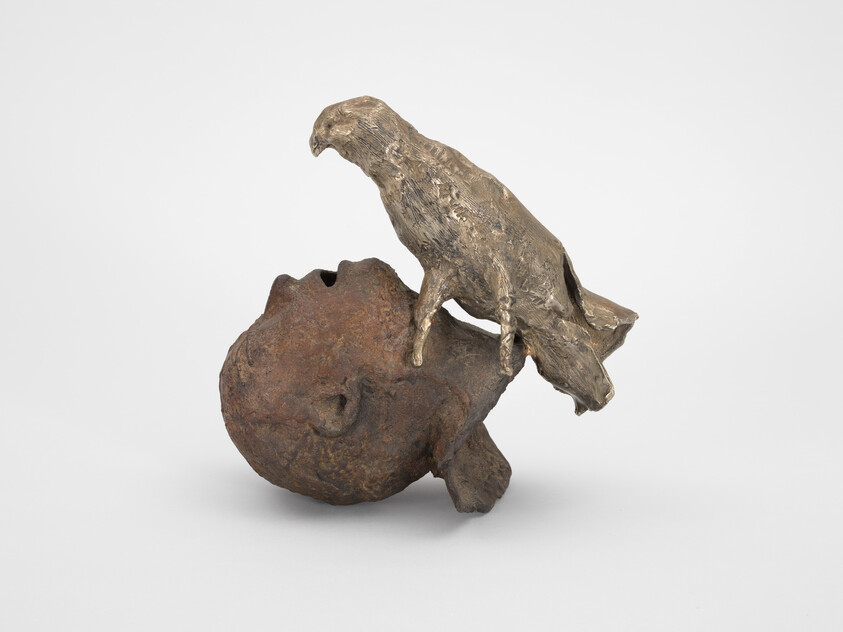 Bronze bird perched on a weathered human skull sculpture, looking downward.